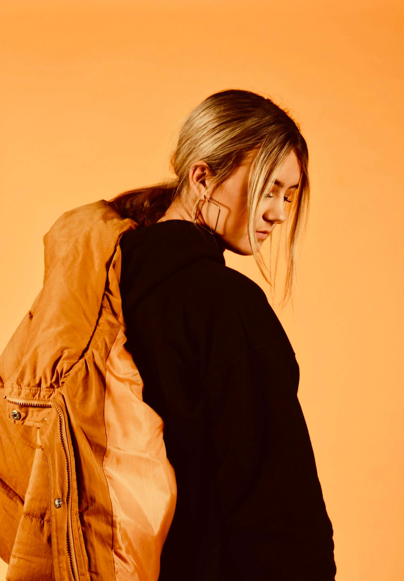 Portrait of a woman in a yellow jacket, posing thoughtfully against a warm orange backdrop.